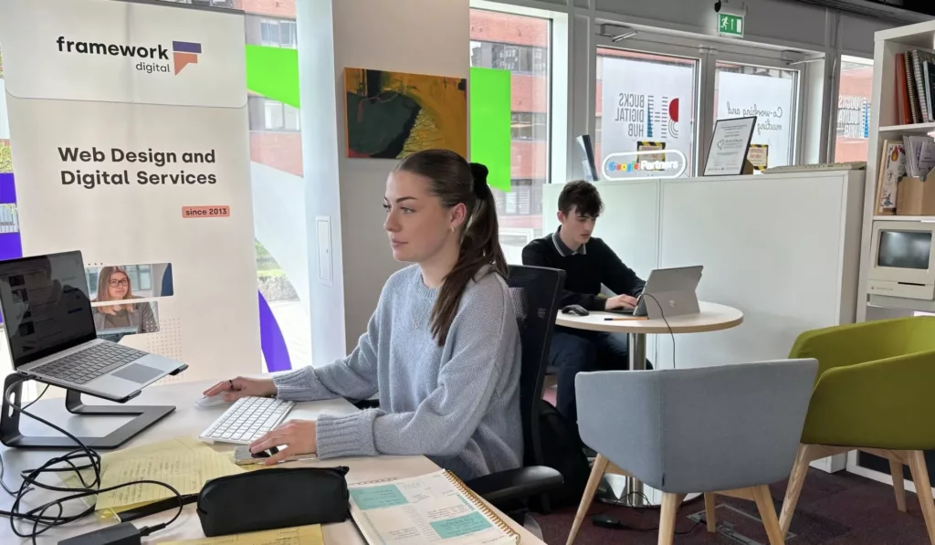 In an office setting a girl is sitting at her desk looking at a computer surrounded by work materials. In the background a young boy is sitting at a table working on a computer.